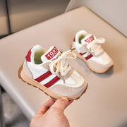 A hand holds a white kids' sneaker with beige suede, red stripes, and a brown sole.